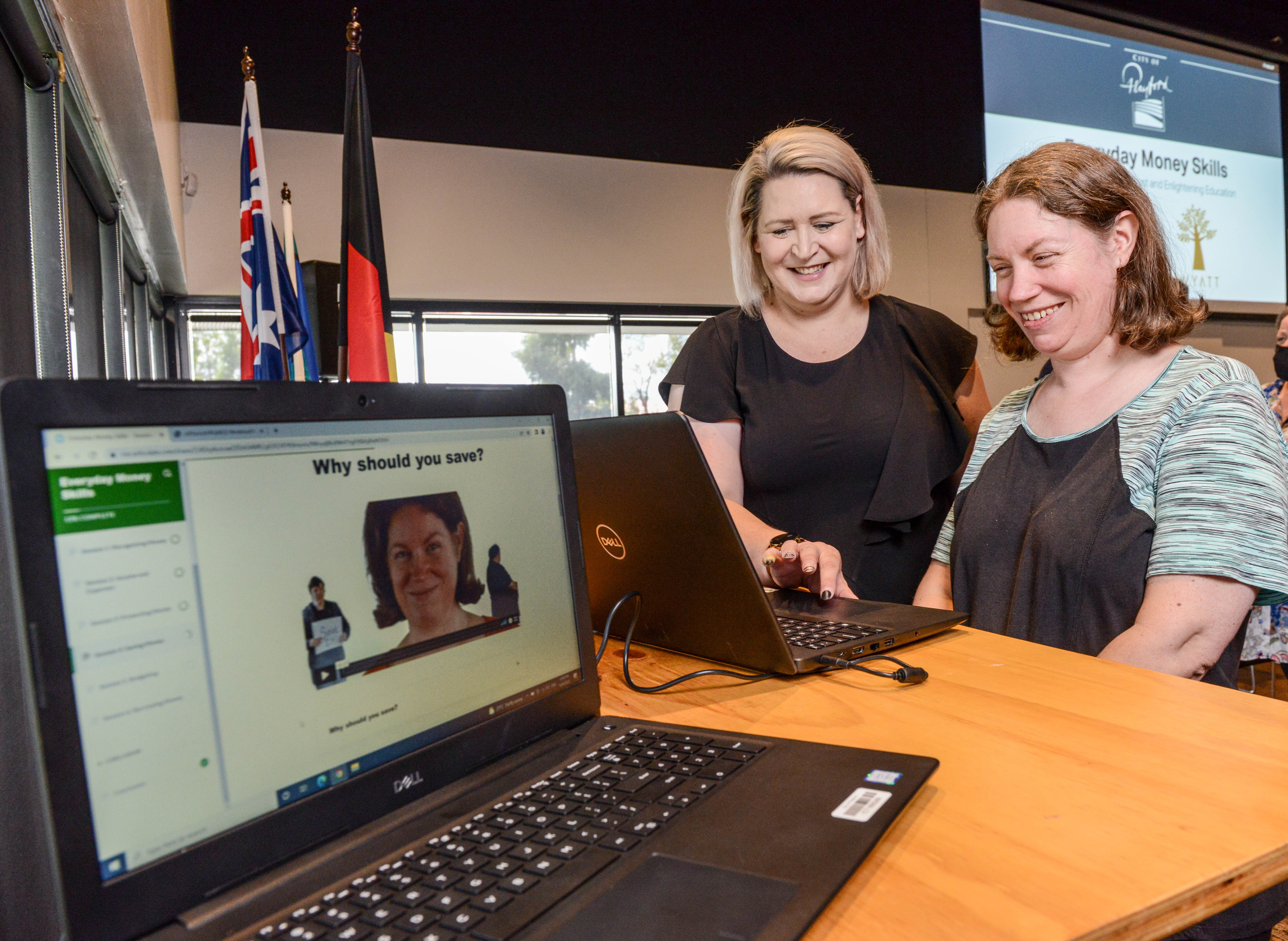 A woman happily shows a person with disability that they’re featured on the everyday money living skills program on a computer. Colourful flags hang in the background.